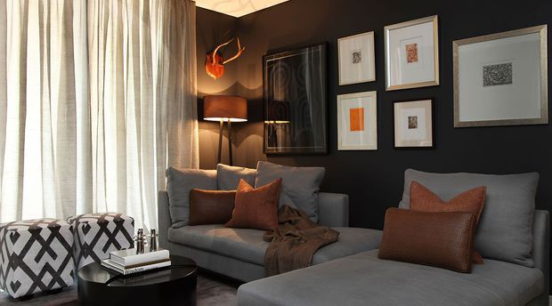 A moody, refined living room with charcoal walls, full-height sheer curtains, a grey sofa and chaise lounge, rust-orange cushions, a dark round coffee table, floor lamp, sculptural wall light, and a curated arrangement of framed artwork. Bold patterned ottomans add graphic contrast.