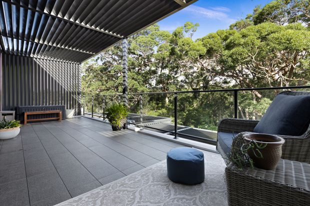 A spacious covered outdoor terrace overlooking dense greenery, with dark tiled flooring, a black metal-framed glass balustrade, a slatted pergola roof, corrugated metal wall cladding, and wicker lounge furniture. Potted plants and a low timber bench add warmth to the contemporary outdoor setting.