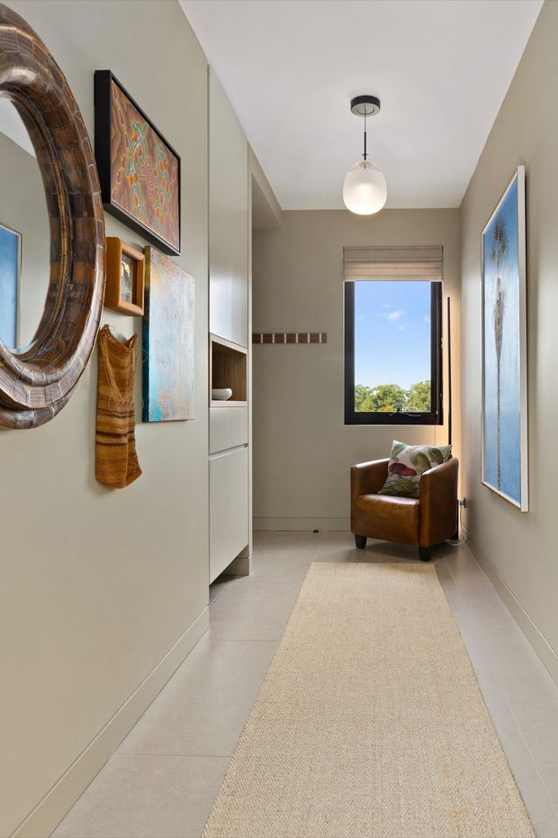 A narrow, softly lit residential hallway with pale greige walls, large light-toned tiled flooring, and built-in cabinetry with open shelving along one side. A long natural-fibre runner leads toward a black-framed window, with a tan leather armchair positioned beneath it. Layered artwork, a large rounded mirror, woven wall hanging, and a simple globe pendant create a warm, curated contemporary interior.