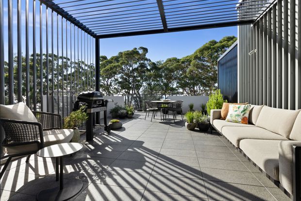 A bright contemporary outdoor terrace with a large tiled entertaining area, covered by a black metal pergola with closely spaced slatted screening. The terrace includes outdoor lounge seating, armchairs, a small coffee table, barbecue, dining table and chairs, and potted greenery. Corrugated metal-clad walls frame the space, with mature trees visible beyond.