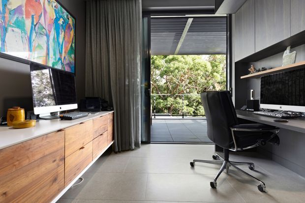 A refined contemporary home office with two workstations. The room features a long timber drawer cabinet supporting a large monitor, a dark built-in desk and shelving wall with a second monitor, grey curtains, large-format light grey floor tiles, and an opening onto a covered balcony or terrace surrounded by greenery. A colourful abstract artwork adds visual interest above the cabinetry.
