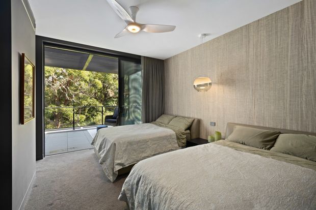 A contemporary bedroom with two beds, soft neutral bedding, a textured vertical wall finish, carpeted flooring, and dark-framed floor-to-ceiling glazing opening onto a balcony or exterior view. A ceiling fan and wall-mounted spherical light add functional and modern details, while the muted palette creates a calm, understated atmosphere.
