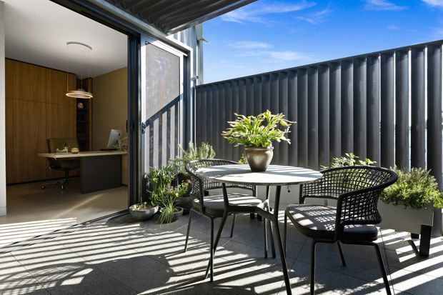 A bright outdoor terrace with a black metal dining table and woven chairs, potted plants, and strong linear shadows cast across grey paving. The terrace is enclosed by a dark corrugated metal privacy screen and opens directly into a compact, warm timber-lined home office with built-in cabinetry and a desk. A retractable or overhead awning provides shelter.