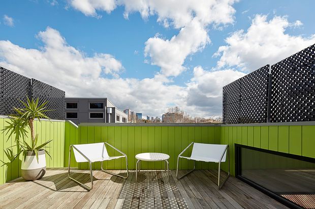 A sunny elevated outdoor terrace with pale timber decking, bright lime-green privacy walls, and black perforated screening panels. Two white sling chairs and a small round table create a compact seating area, with a potted architectural plant at the edge. Modern low-rise buildings and a blue, cloud-filled sky are visible beyond the terrace.