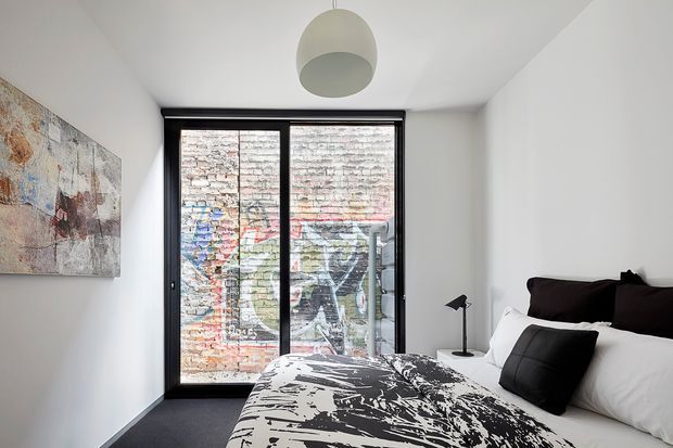 Compact contemporary bedroom with white walls and ceiling, dark carpet, a low-profile bed dressed in white linens and a bold black-and-white graphic duvet, and black accent pillows. A large black-framed glazed door or window opens the room visually to a colourful brick graffiti wall outside. Minimal furnishings include a small black bedside lamp, a large abstract artwork, and a simple round pendant light.