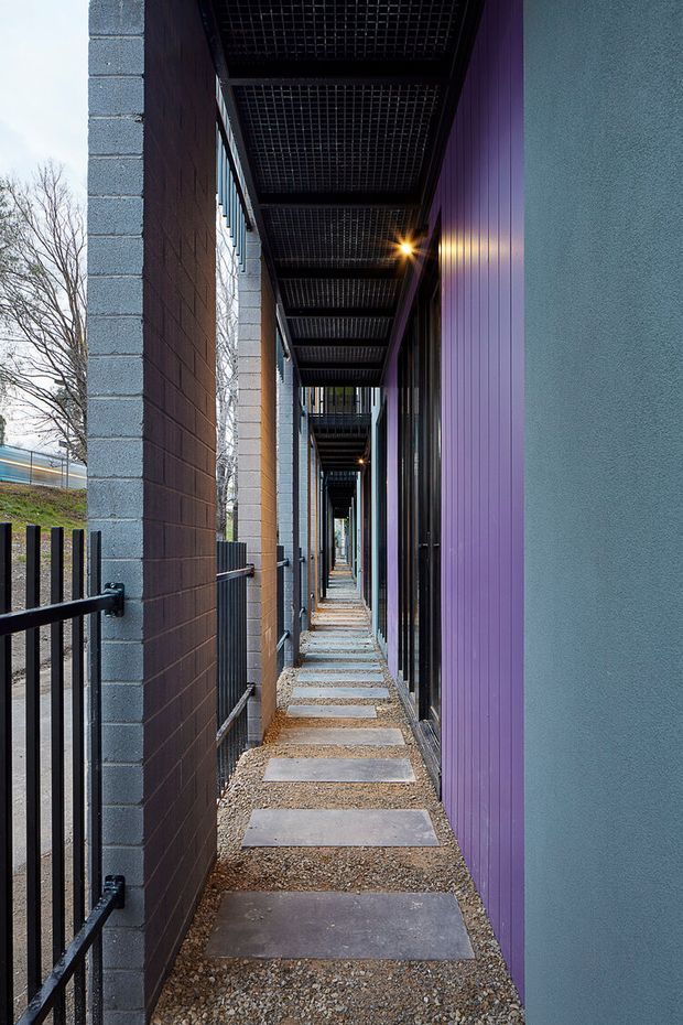 A narrow covered external walkway between modern building wings, with repeating dark steel overhead grating, exposed blockwork columns and walls, vertical purple cladding, glazed openings, black metal railings, and a gravel path with rectangular concrete stepping slabs. The strong linear perspective emphasizes the corridor-like circulation space.