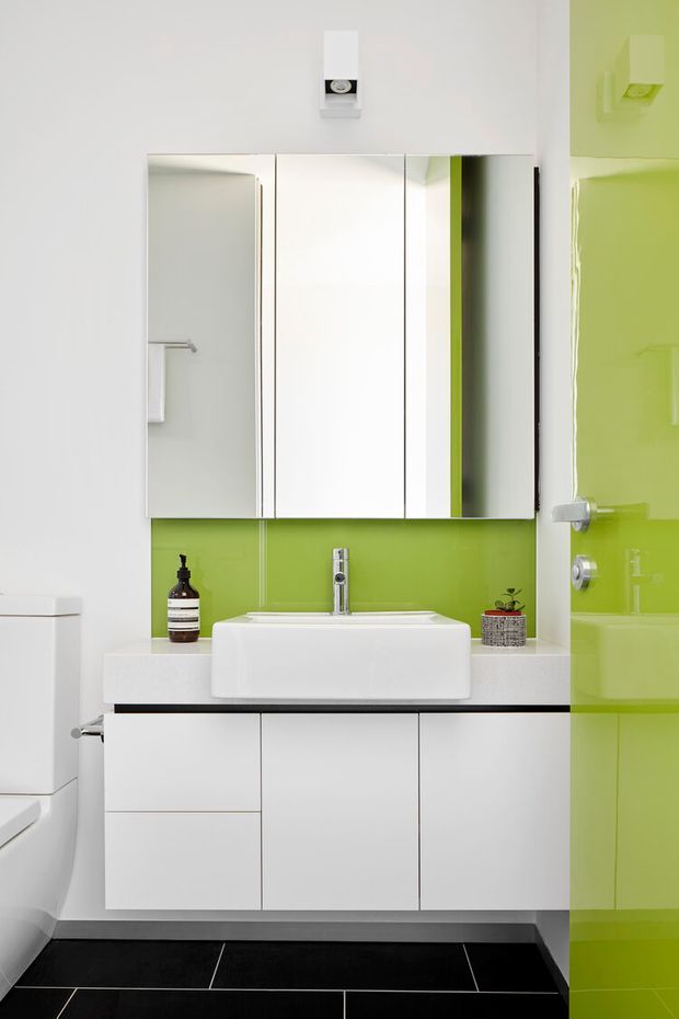 Compact contemporary bathroom with a wall-mounted white vanity, rectangular vessel basin, mirrored cabinet, vivid lime-green glass wall and door, and dark tiled floor. A toilet is partially visible at left, with minimal toiletries and a small plant on the vanity.