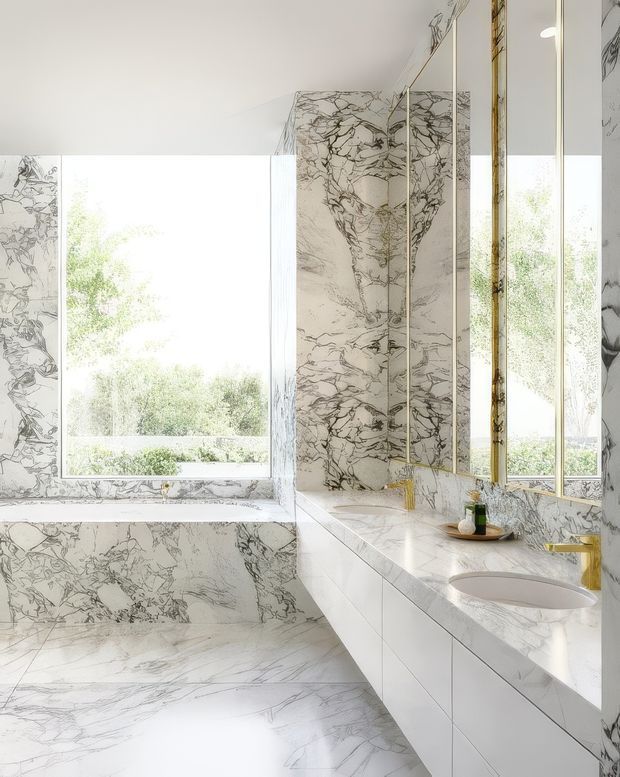 Bright, luxurious bathroom clad extensively in white marble with dramatic grey veining. The room features a large built-in marble bathtub beneath a wide window, a long white vanity with integrated basins and drawers, tall frameless mirrors with slim metallic trim, and brushed gold tapware and accessories. Large windows bring in abundant natural light and views of greenery, creating an airy, high-end atmosphere.