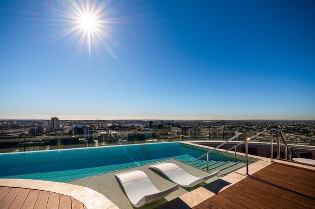 Rooftop or elevated infinity pool overlooking a dense coastal cityscape, with turquoise water, integrated curved pool loungers, stainless-steel handrails, and warm timber decking under a bright blue sky.