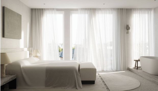 Bright, serene contemporary bedroom with a low upholstered bed, neutral bedding, a pale upholstered bench, sheer floor-to-ceiling curtains, and abundant natural light. A freestanding bathtub is visible at the right, suggesting an open bedroom-bathroom arrangement. The room uses a restrained palette of white, cream, and light grey with minimal decor and soft, refined finishes.