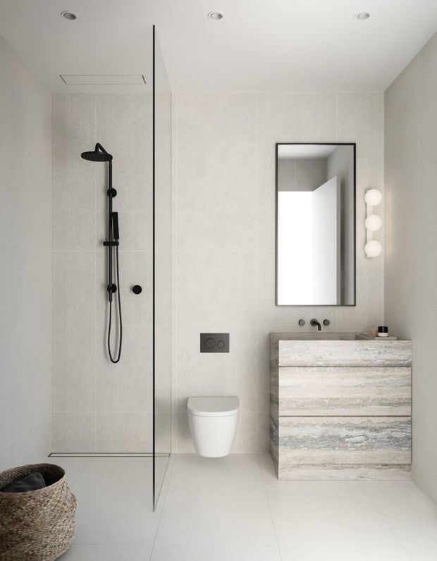 A contemporary minimalist bathroom with a walk-in shower enclosed by a frameless glass screen, wall-mounted black shower fittings, a wall-hung toilet, and a floating textured stone-look vanity with integrated basin. A black-framed rectangular mirror and vertical globe wall lights complement the pale tiled surfaces and neutral palette.
