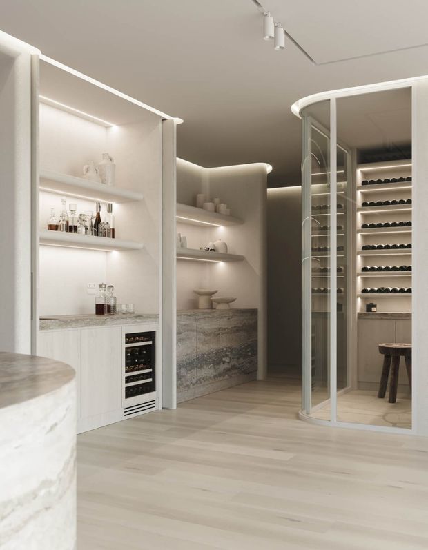 A refined, softly illuminated home bar and wine storage area with curved glass wine-room doors, extensive bottle racks, built-in shelving, integrated refrigeration, and a pale stone bar counter. The space uses warm concealed lighting, light timber flooring, and pale neutral finishes for a luxurious contemporary feel.