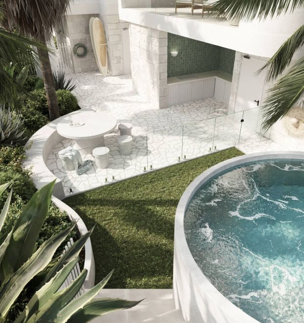 A bright tropical residential courtyard viewed from above, featuring a curved raised swimming pool with turquoise water, a white stone-paved outdoor entertaining area, circular dining furniture, glass pool fencing, lush palm and agave planting, and a built-in outdoor bar or kitchenette beneath a white upper level. The architecture has a clean contemporary coastal character with pale masonry and a green tiled recess.