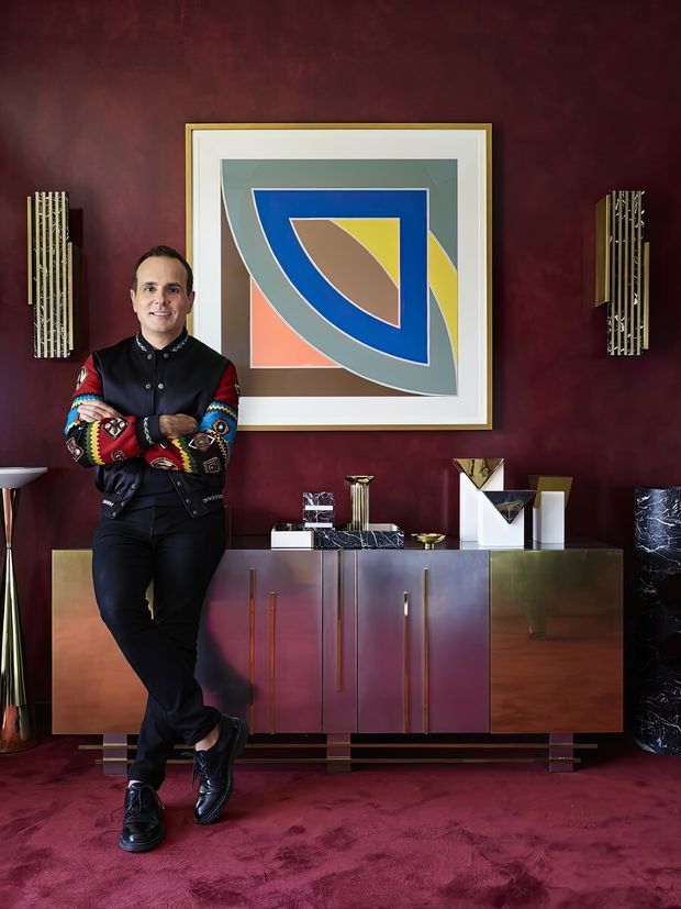 A richly styled interior portrait showing a person standing beside a sculptural, multicoloured sideboard against a deep burgundy wall. The room features a large geometric abstract artwork in a pale frame, metallic cabinet fronts and hardware, decorative objects, fluted wall sconces, and a plush burgundy carpet. The composition has a highly curated, glamorous, art-forward character with strong geometric forms and saturated colour.