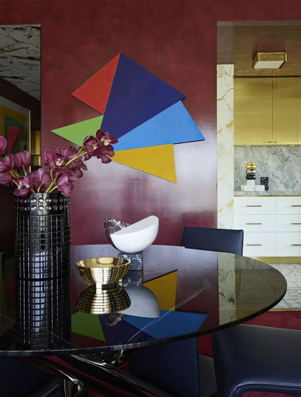 A richly styled dining area with a glossy dark oval dining table, sculptural chairs, a tall black vase holding purple orchids, and metallic decorative objects. The deep burgundy wall features a bold geometric multicolour artwork, while a marble-framed kitchen with pale cabinetry is visible through an opening at right. The interior has a dramatic, luxurious Art Deco-inspired character with strong reflective surfaces and saturated colours.