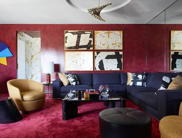 A richly styled living room with deep burgundy walls and matching plush carpet, a large dark navy sectional sofa, sculptural mustard armchairs, and a black leather ottoman. Abstract geometric artworks are arranged on the feature wall, with a mirrored panel on the right and a distinctive contemporary pendant light overhead. The room has a bold, luxurious, high-contrast aesthetic with strong mid-century and Art Deco influences.