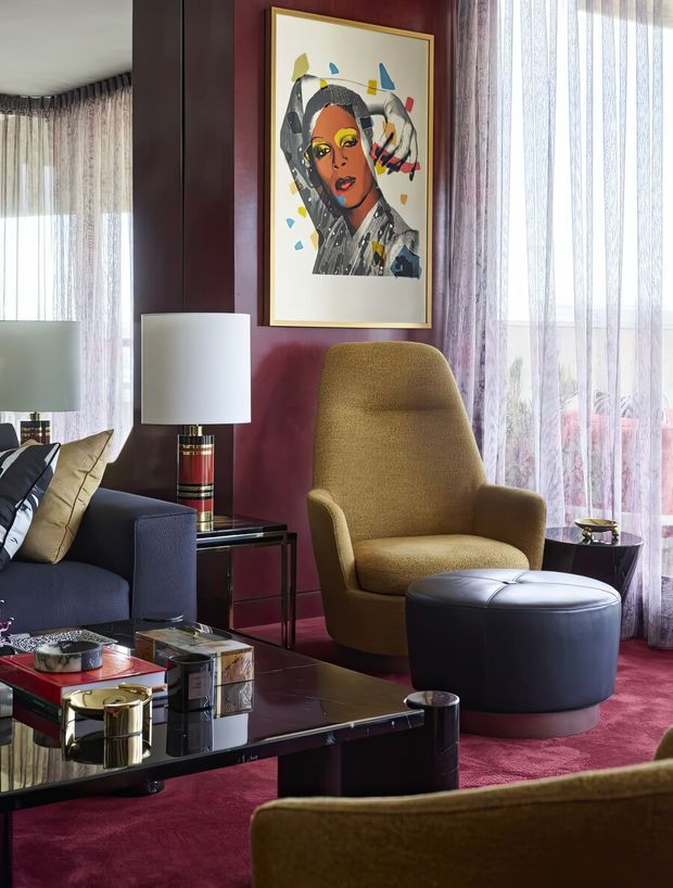 A richly layered living room with deep burgundy walls and carpet, full-height sheer curtains, a dark blue sofa, mustard upholstered lounge chair, and matching ottoman. A glossy black coffee table, sculptural lamps, framed artwork, and reflective surfaces create a sophisticated, colourful interior with strong geometric and vintage-inspired detailing.