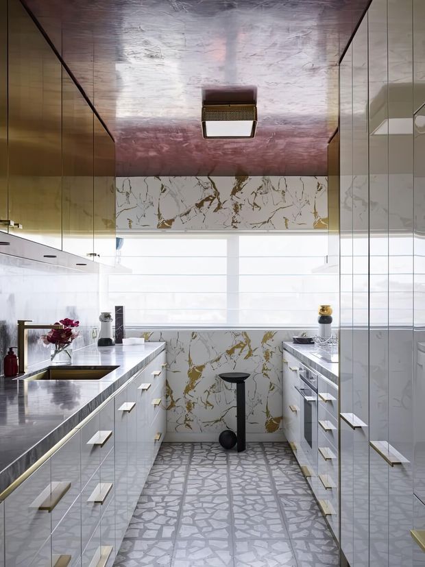 A narrow, highly polished galley kitchen with extensive glossy white cabinetry, long stone-look benchtops, integrated appliances, and a dramatic white marble-look splashback with bold gold veining. Reflective metallic wall and ceiling finishes add a luxurious, contemporary character, while a large bright window illuminates the space. A patterned stone-look floor runs between the parallel banks of cabinetry.