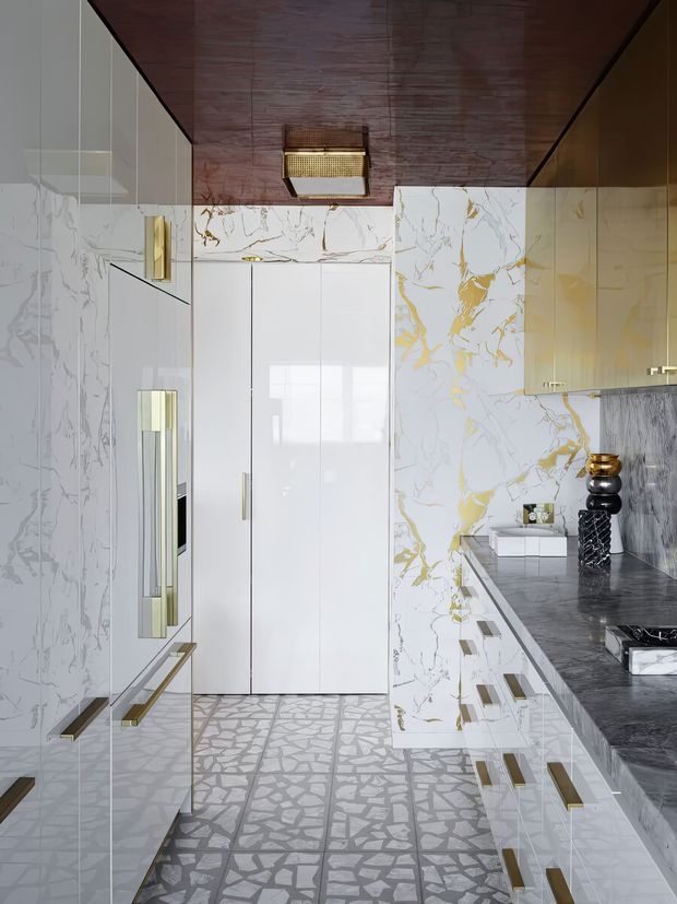 A luxurious contemporary kitchen viewed down a narrow galley-like space. Glossy white full-height cabinetry and marble-look wall panels with dramatic grey veining and gold accents are paired with a dark grey stone benchtop and matching dark timber ceiling. The floor features a striking irregular grey-and-white patterned tile, while brass-and-gold hardware, integrated appliances, and decorative lighting add refined contrast.