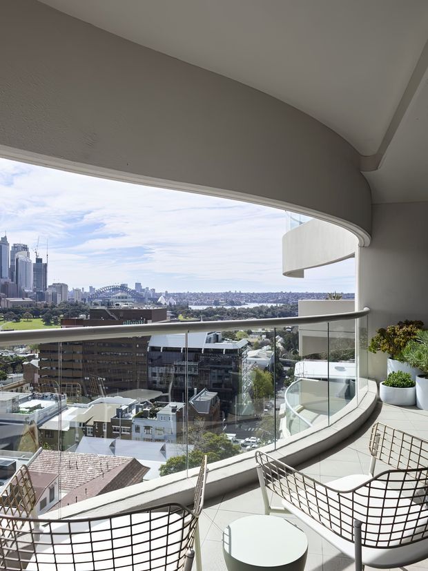 A bright, elevated balcony or terrace with a sweeping curved concrete soffit and balustrade, furnished with contemporary wire-frame chairs and a small round table. Glass railings provide expansive views over a dense urban neighbourhood, harbour, and prominent city skyline landmarks, including a large arched bridge. Potted plants add greenery along the curved edge.