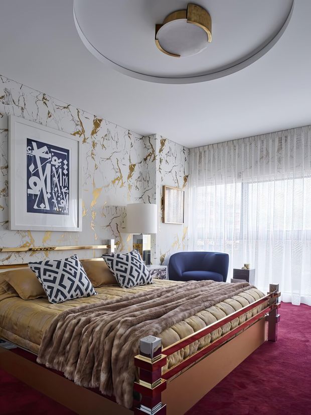 A glamorous bedroom with a large upholstered bed featuring dark red and brass detailing, layered taupe bedding, geometric patterned cushions, and a rich burgundy carpet. The walls are covered in white marble-look wallpaper with bold gold veining, complemented by modern graphic artwork, a sculptural ceiling light, a floor lamp, a blue lounge chair, and full-height sheer window blinds. The room has a luxurious, dramatic, art-deco-inspired character.
