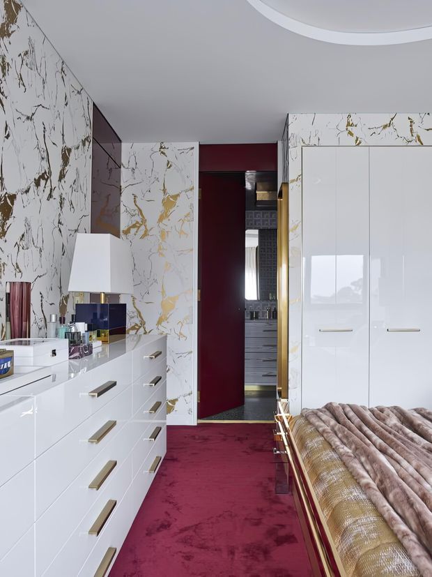 A luxurious bedroom dressing area with glossy white built-in drawers and wardrobes, dramatic white marble-effect wall panels with gold veining, a deep burgundy wall and matching plush carpet, and a partially visible bed with a metallic frame and textured fur throw. A doorway leads toward a darker bathroom or dressing space. The room has a refined, high-contrast, glamorous aesthetic with a curved recessed ceiling detail.