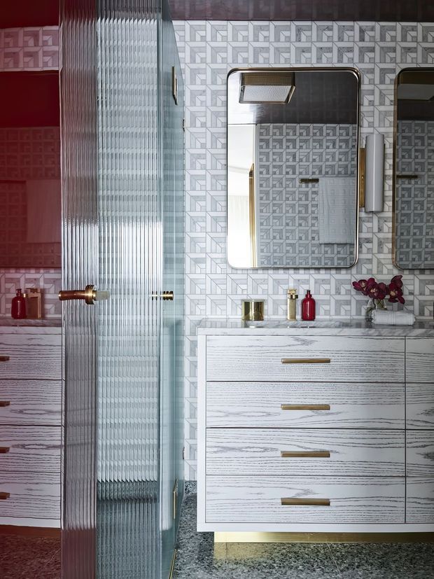 A stylish bathroom featuring a white-and-grey geometric patterned tile wall, a large floating white wood-grain vanity with brushed gold handles, integrated countertop basin, and twin mirrored cabinets. A ribbed textured glass shower enclosure divides the vanity from the shower area. Burgundy-red cabinetry and accessories, gold fixtures, dark patterned floor tiles, and orchid décor create a bold, luxurious, art-deco-inspired scheme.