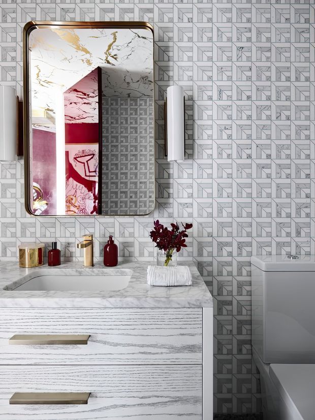 A highly styled contemporary bathroom vanity with a white marble-effect benchtop and integrated basin, paired with a timber-grain vanity featuring long brushed-gold handles. The walls are covered in a geometric grey-and-white patterned tile, and a rounded rectangular mirror with a bronze-gold frame hangs above the vanity. Gold and deep burgundy accessories, a white wall sconce, folded towels, and a compact toilet complete the refined, graphic interior.