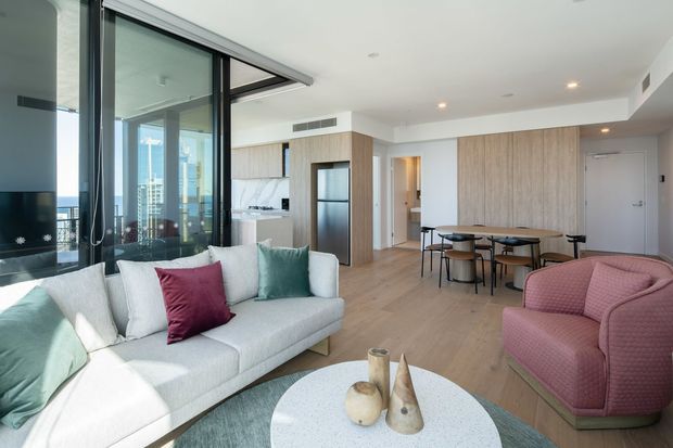 Bright open-plan apartment interior combining a living room, dining area and compact kitchen. Floor-to-ceiling glazing provides expansive city views, while pale timber flooring and cabinetry create a light, contemporary setting. The room is furnished with a light grey sectional sofa, pink lounge chairs, a round dining table, and colourful cushions.