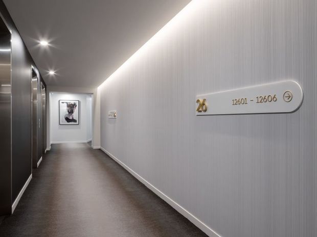 A sleek, contemporary hotel or commercial corridor with dark polished flooring, subtly textured light wall panels, recessed ceiling lighting, and dark elevator or door panels along the left side. A directional room-number sign with metallic gold lettering is mounted on the right wall, and framed artwork marks the end of the hallway.