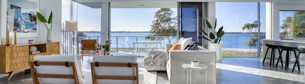 Bright open-plan coastal living space with expansive floor-to-ceiling glazing overlooking a calm waterfront. The room includes a light upholstered sofa, timber-framed lounge chairs, a timber sideboard, indoor plants, and a compact kitchen or dining area with bar seating. Pale finishes and abundant daylight create an airy, relaxed atmosphere.