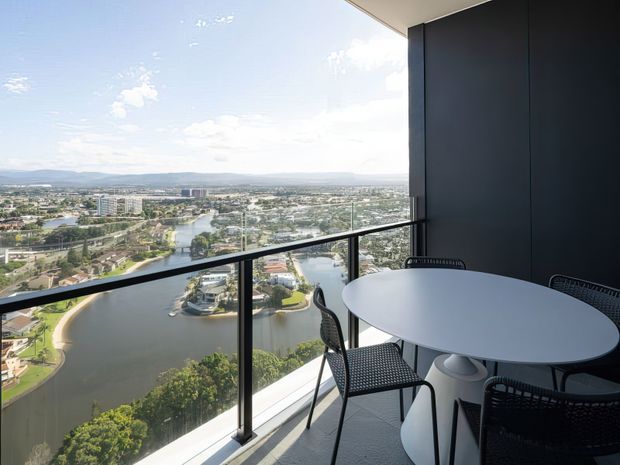 High-rise balcony or terrace with floor-to-ceiling glazing and a glass balustrade, furnished with a round white table and dark woven chairs. The space overlooks a broad river or lake, surrounding urban development, and distant hills in bright daylight.