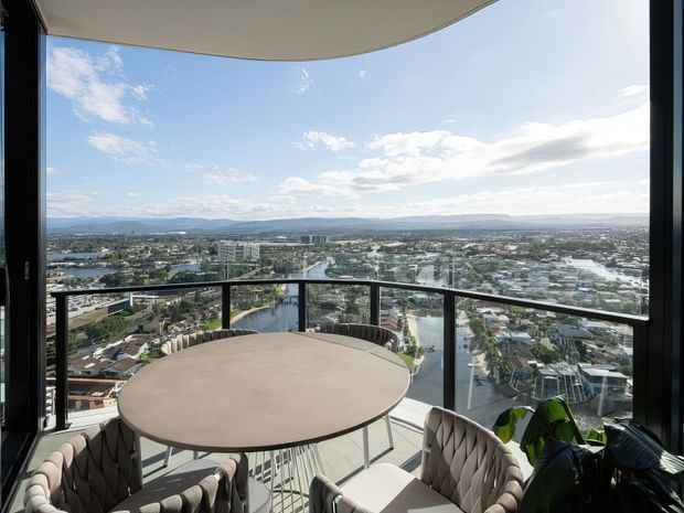 A high-rise balcony or terrace with floor-to-ceiling glazing and a glass balustrade, furnished with a round dining table and upholstered chairs. The space overlooks a dense urban area with waterways and distant hills under a bright sky. A curved projecting ceiling or soffit frames the terrace.