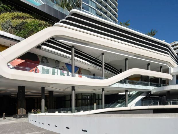 Exterior view of a large contemporary multi-storey commercial or mixed-use building with sweeping curved white balconies and layered horizontal rooflines. The facade combines glass balustrades, dark slender columns, extensive glazing, integrated greenery, and an external escalator, with a mural visible inside the open frontage. The image is captured from an outdoor plaza under a clear blue sky.