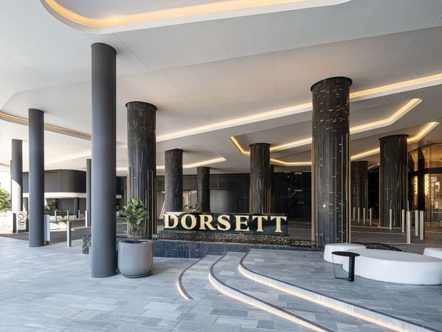 A sophisticated hotel entrance or lobby featuring a prominent DORSETT sign, tall dark cylindrical columns, black textured feature walls, curved tiered steps, integrated warm cove lighting, and pale grey stone-look paving. The space has a polished contemporary hospitality design with a dramatic yet refined atmosphere.
