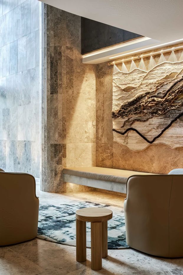 A refined contemporary interior featuring two rounded upholstered lounge chairs facing a sculptural textured wall artwork. The space has warm stone wall and floor surfaces, a built-in upholstered bench, a patterned area rug, a small timber side table, and concealed linear lighting beneath a layered ceiling detail. A large glazed opening brings in soft natural light.