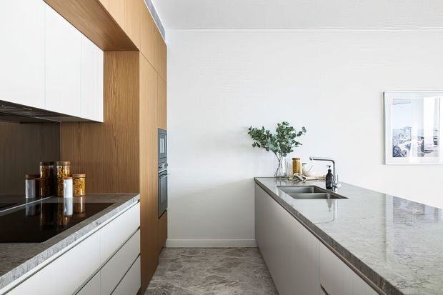 A bright contemporary kitchen with a long central island, integrated sink and tap, white cabinetry, a dark stone cooking surface, and a tall warm timber joinery wall enclosing built-in appliances. The room has pale walls, a grey stone-look floor, and restrained styling with a small vase and framed artwork.