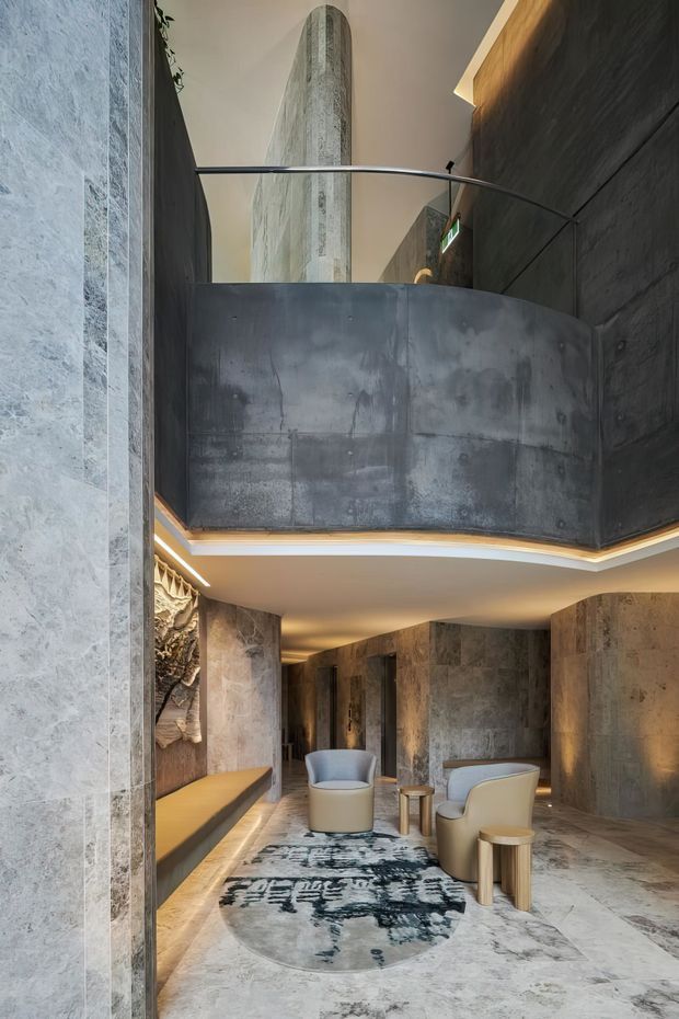 A dramatic multi-level interior lounge or entry space with curved concrete and dark stone walls, a sweeping blackened metal mezzanine balustrade, softly integrated cove lighting, sculptural upholstered lounge chairs, and a large abstract rug. A tall cylindrical textured architectural feature rises through the upper level, creating a sculptural focal point.