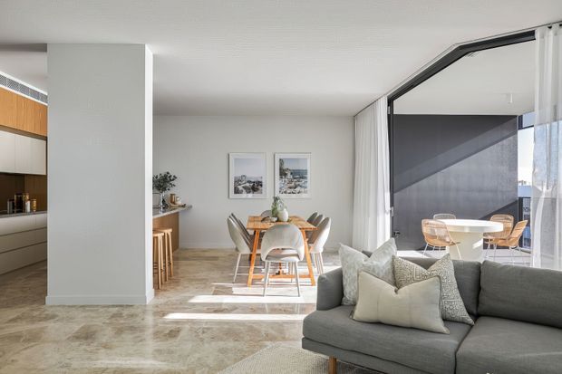 Bright contemporary open-plan apartment interior with a living room in the foreground, dining area centrally positioned, and a partially visible kitchen at left. The space features pale stone-look flooring, white walls and ceiling, timber kitchen cabinetry, a timber dining table with upholstered chairs, a grey sectional sofa, and expansive black-framed glazing opening to an outdoor terrace or balcony. Natural light creates a calm, airy atmosphere.