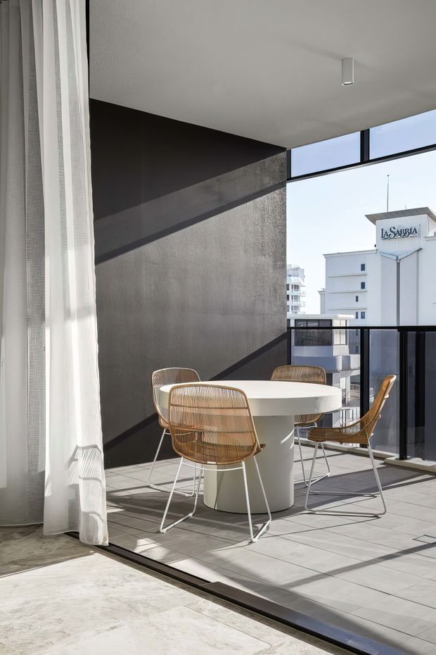 Bright contemporary dining area opening onto a balcony or terrace, with a round white pedestal table and four woven cane dining chairs. Full-height sheer curtains, dark textured feature walls, pale tiled flooring, black-framed glazing and balustrades, and strong natural light create an airy urban apartment setting. A neighbouring building is visible beyond the glazing.