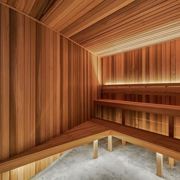 A compact sauna lined throughout with warm-toned vertical timber wall panels and horizontal timber ceiling slats. Tiered built-in timber benches wrap around the room, with concealed linear lighting beneath the upper bench and a pale stone or concrete floor below.