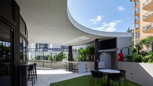 A contemporary rooftop terrace or courtyard lounge with a sweeping curved canopy, dark metal-framed glazing and vertical screening, pale stone paving, built-in bar seating, and outdoor café-style furniture. The space is bright and open, with adjacent multi-storey buildings visible beyond.