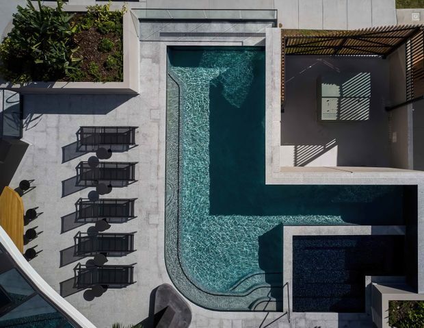 Overhead architectural photograph of a contemporary outdoor pool courtyard. A geometric swimming pool with a curved edge, shallow entry or sun shelf, and darker inset section occupies the center, surrounded by pale stone or concrete paving. Lounge chairs and small round tables line the pool, with a planted garden bed at the upper left. A shaded pergola or louvred outdoor structure sits beside the pool, casting strong linear shadows.