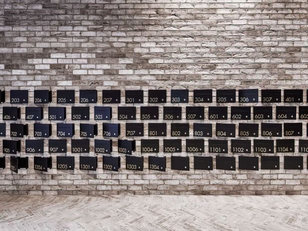 A corridor or lobby feature wall clad in exposed grey-white brick, covered with rows of black numbered apartment-style mailboxes. The floor has a pale herringbone or parquet-like pattern, creating an industrial, utilitarian building entrance aesthetic.
