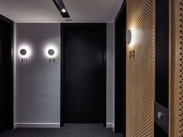 A dimly lit contemporary hallway or lift lobby with dark carpet, matte black doors, grey plaster walls, circular wall lights, room numbering, and a prominent perforated timber screen on the right. The space has a refined, moody hotel or apartment-building character.