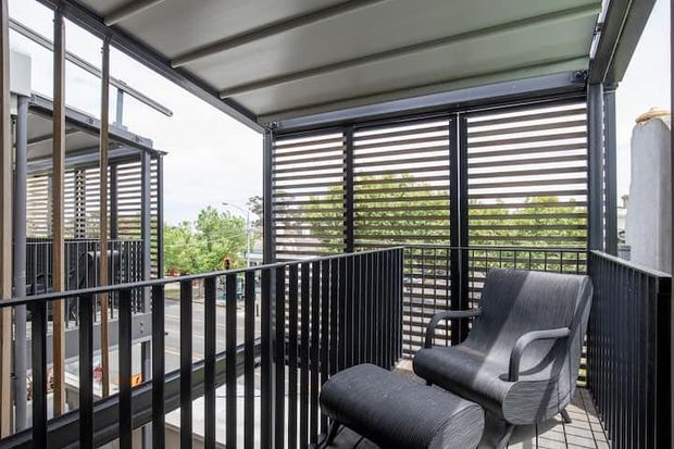 A compact elevated outdoor balcony or terrace with dark metal balustrades, a covered flat roof, and horizontal louvred privacy screens. The space contains a dark upholstered lounge chair with matching footstool and overlooks leafy surroundings; neighbouring balconies or terraces are visible to the left.