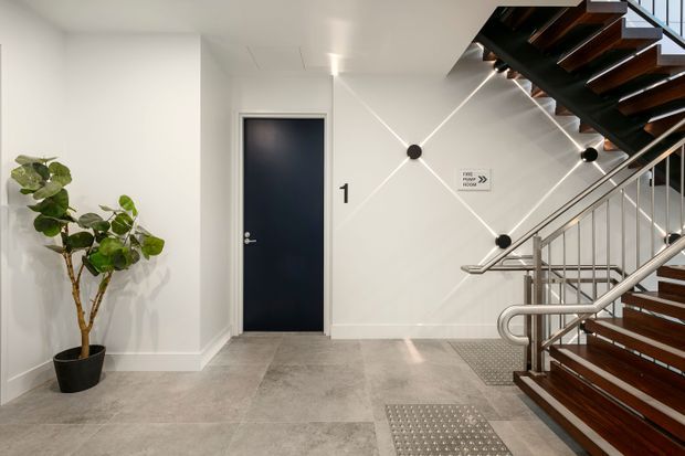 A bright contemporary stairwell or building landing with white walls, grey tiled flooring, a dark navy door, stainless-steel handrails, and an open stair with dark timber treads. Linear wall lighting creates a geometric pattern, while tactile floor indicators and directional signage suggest a public or commercial building. A potted indoor tree adds greenery.