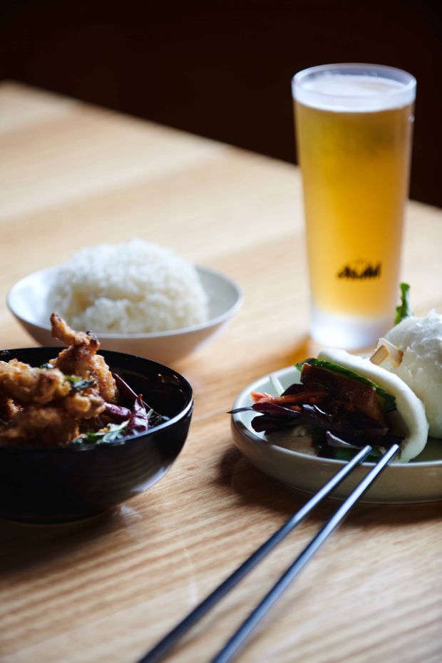 Close-up food photograph on a light timber table, showing bowls of rice and Asian-style dishes with chopsticks, alongside a tall glass of golden beer. The image focuses on dining and food rather than architectural or interior design features.