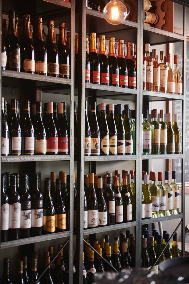 A tall, dark-toned wine display with multiple industrial-style metal shelving bays densely stocked with wine and champagne bottles. Warm pendant lighting illuminates the shelves, while chalkboard-style writing and a partial counter suggest a wine shop, cellar, or bar setting.