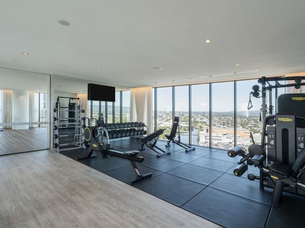 A spacious, modern home gym with extensive strength-training equipment, free weights, benches, and rubber gym flooring. The room has pale timber-look flooring, mirrored walls, a wall-mounted screen, recessed ceiling lights, and full-height glazed windows offering expansive city views.